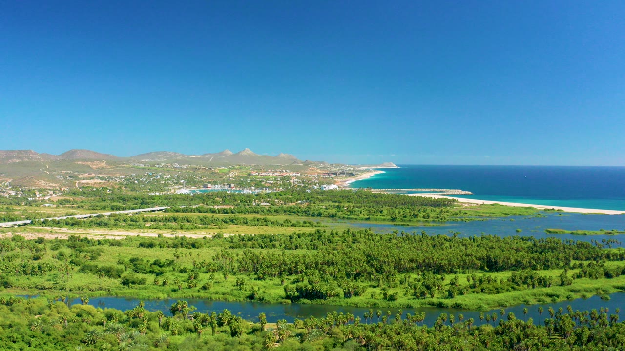 Opening aerial frame over San Jose del Cabo wetlands, with green fields and river water in front, sandy beach and blue ocean to the right, distant hills and no visible people.