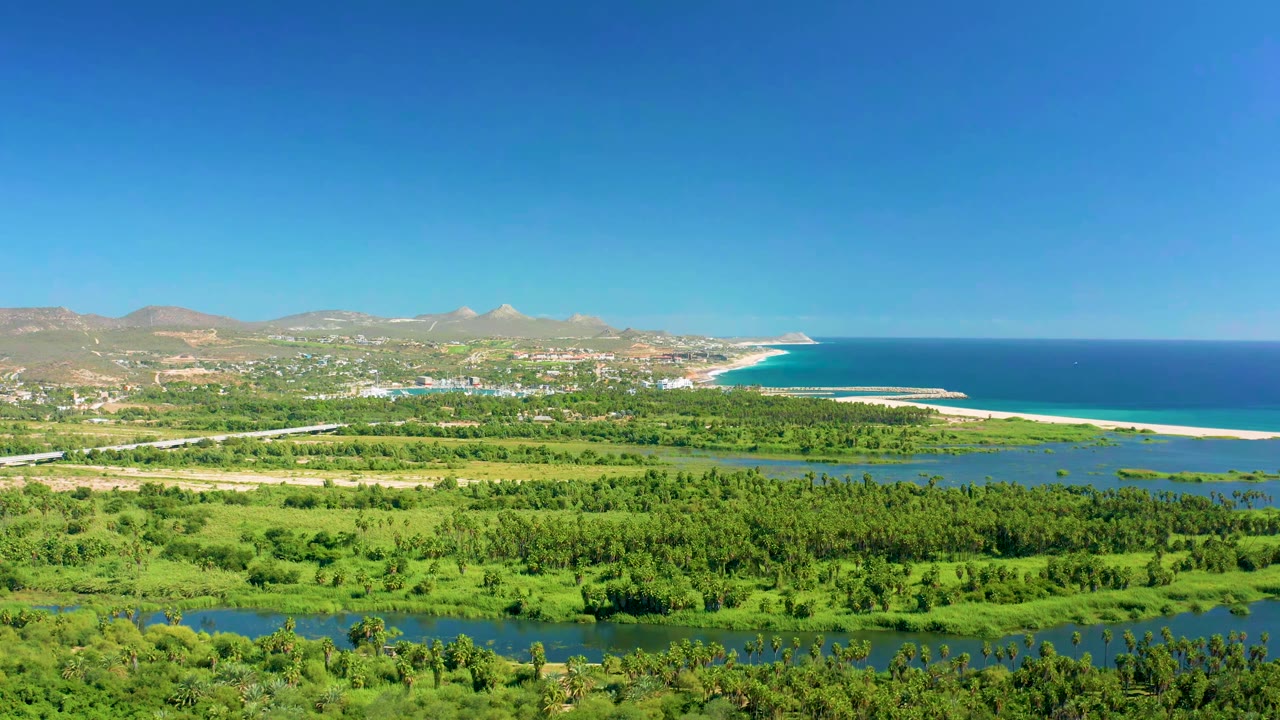 Aerial frame over San Jose del Cabo showing lush green fields, river channels, a pale sand beach, deep blue ocean, distant mountains, coastal buildings, and no visible people.