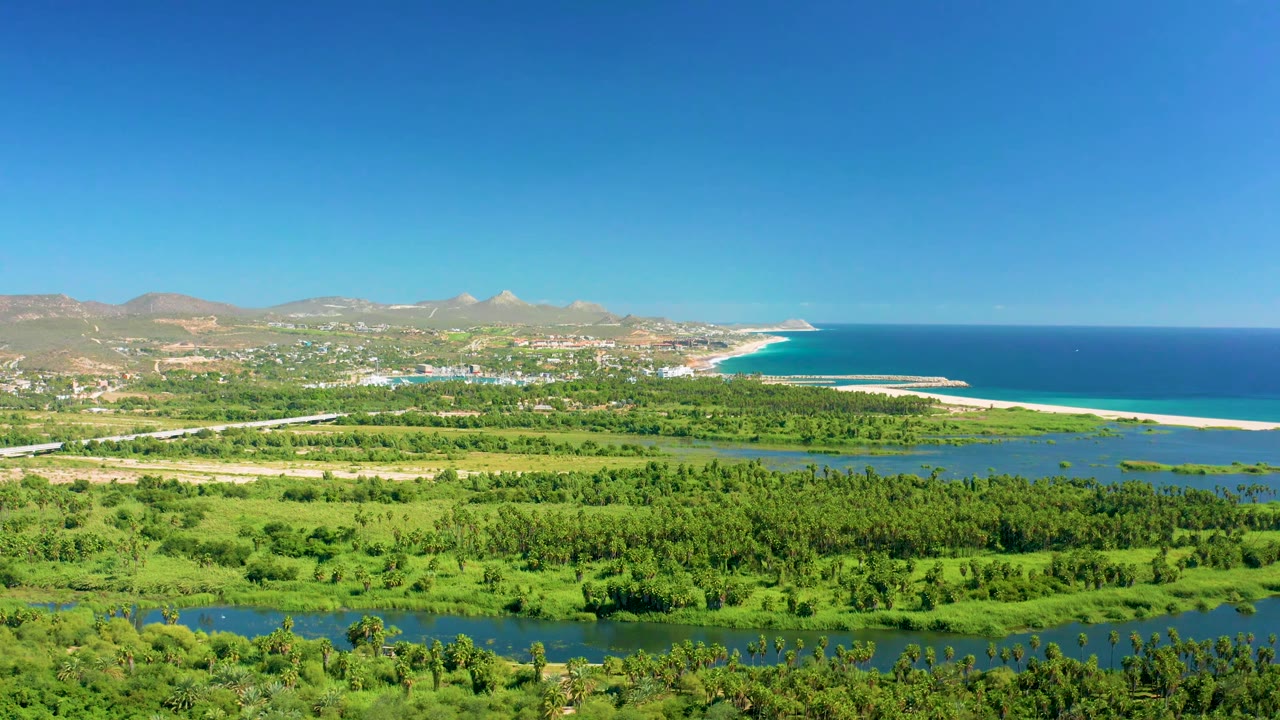 Wide San Jose del Cabo aerial with dense green vegetation below, a river crossing the middle of frame, coastal buildings near the hills, ocean on the right, and no visible people.