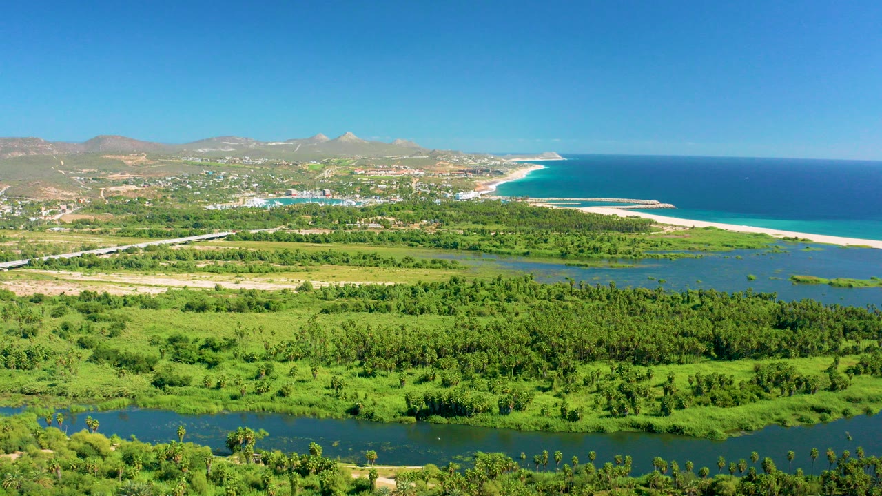Aerial view over San Jose del Cabo with green fields in front, a river channel through the middle, a sandy beach and blue ocean beyond, distant mountains, and no visible people.