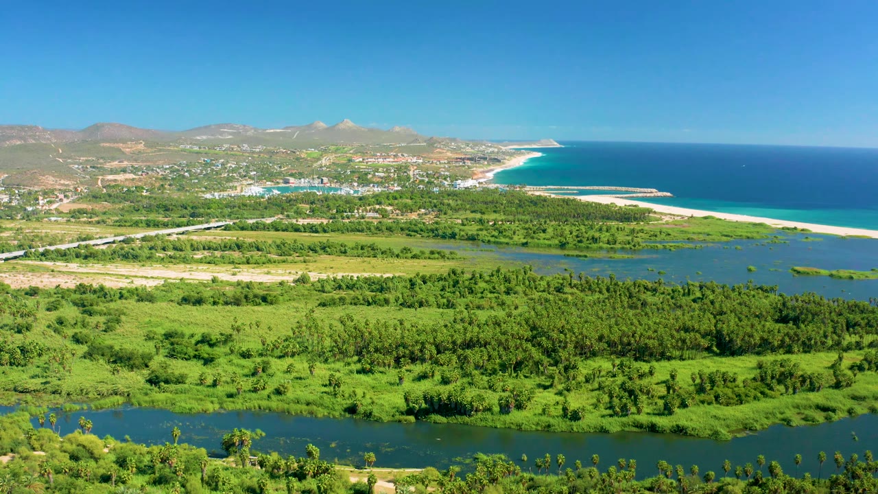 Aerial view in San Jose del Cabo with the river and green fields below, the town and mountains in the distance, and the beach and ocean stretching along the right edge, with no visible people.