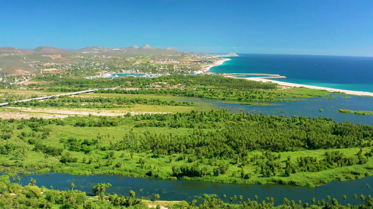 San Jose del Cabo aerial frame showing dense green fields and water channels in the foreground, dry hills and buildings beyond, and a curved beach meeting the blue sea, with no visible people.