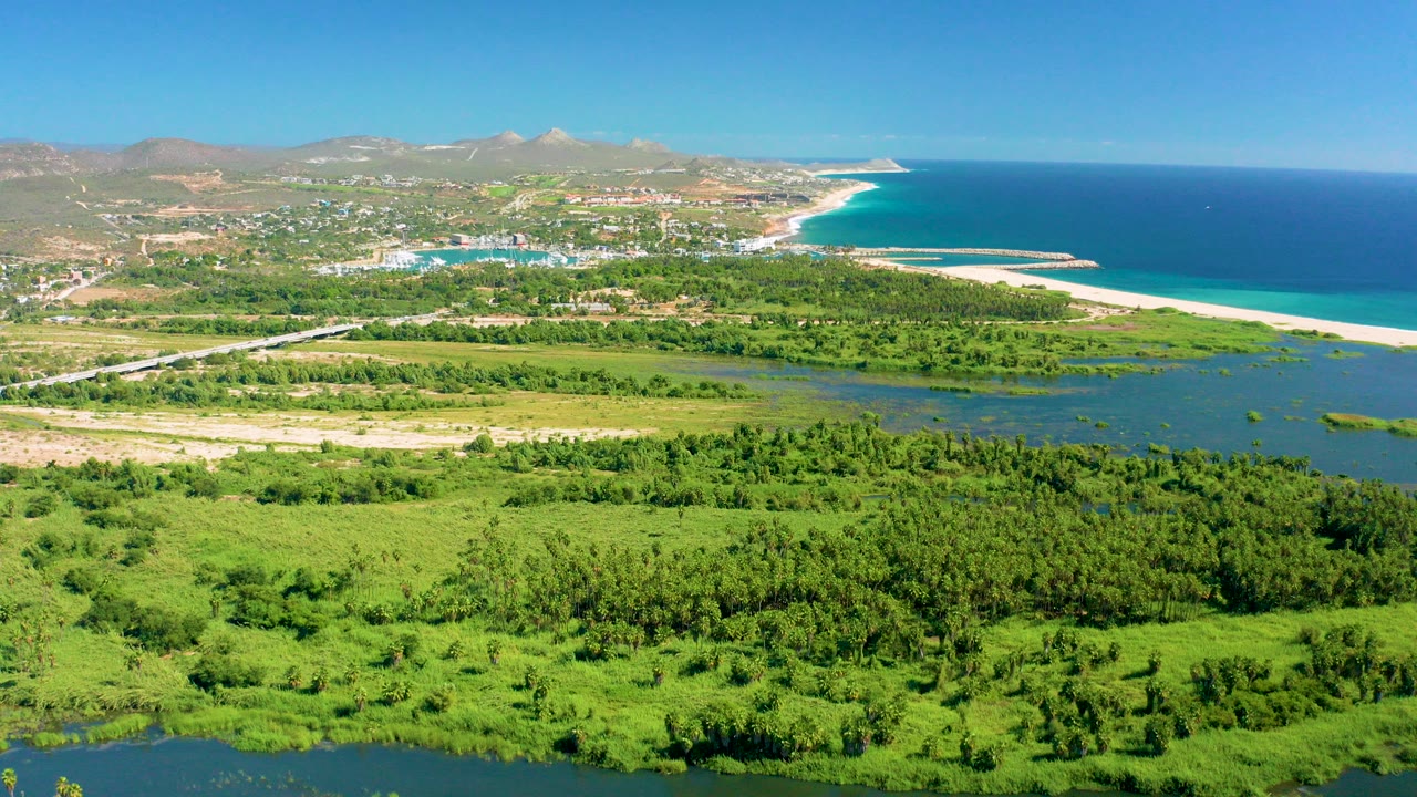 Aerial view over green fields and lagoon water toward a road, sandy beach, blue ocean, and hills in San Jose Del Cabo, with no visible people.