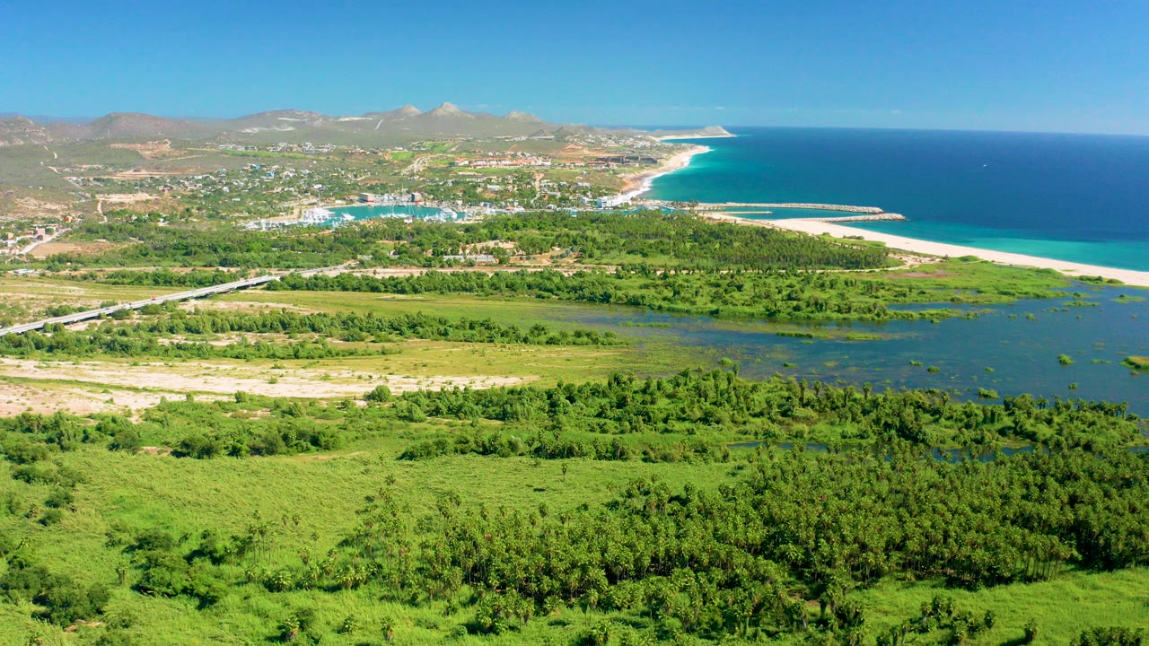 Wide aerial view over San Jose Del Cabo, Baja California Sur, with green fields in front, a lagoon band, sandy beach, blue ocean, distant settlement, mountains, and no visible people.