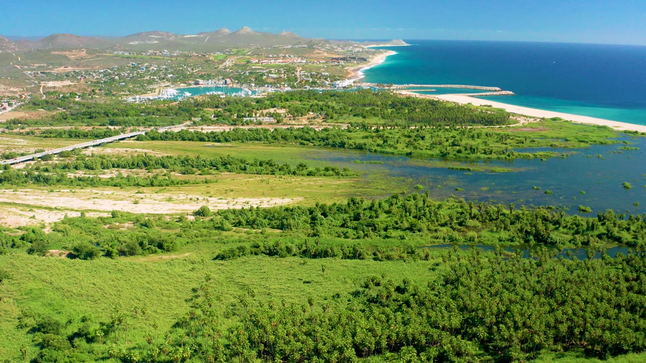 Wide aerial view over San Jose del Cabo wetlands with dense green foreground, a lagoon, sandy beach, blue ocean, distant town, mountains, and no visible people.
