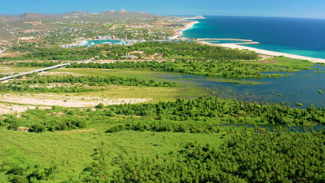 Drone-like view of green coastal marsh, lagoon, beach, ocean, and distant hills.