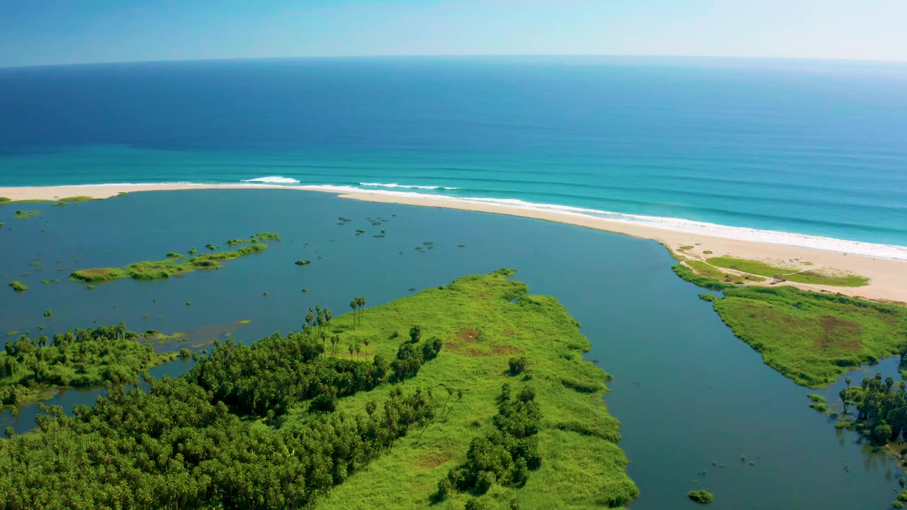 High aerial frame in San Jose del Cabo with the estuary running beside dense green vegetation, pale sand, blue ocean bands, a straight horizon, and no visible people.