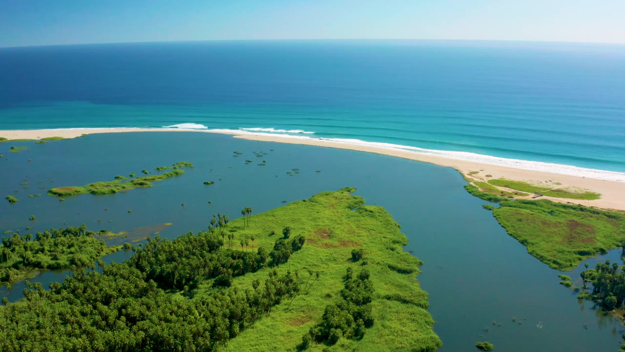 Aerial frame in San Jose del Cabo with a broad estuary mouth, green shoreline vegetation, a pale beach, rolling white surf near the sand, and no visible people.