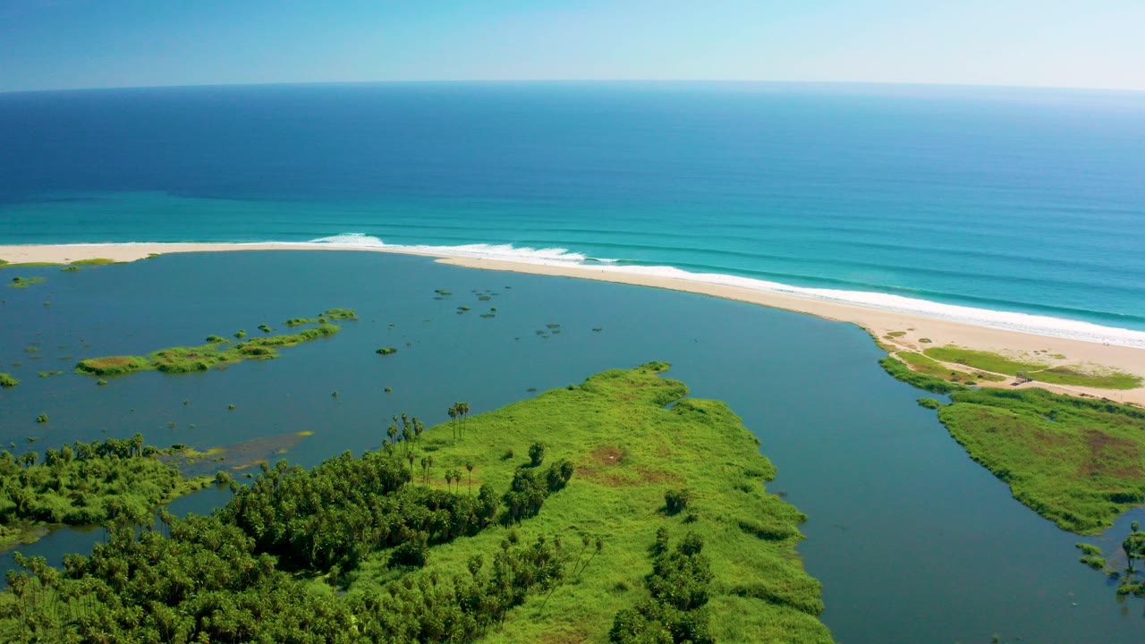 Aerial frame over the San Jose del Cabo estuary with green land in the foreground, blue estuary water, a pale sand beach, white surf, and no visible people.