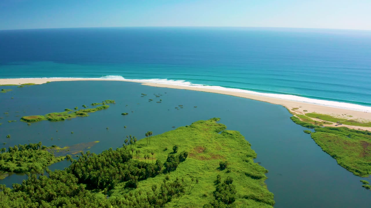 The San Jose del Cabo river estuary appears calm under bright light, with scattered green islands, a long white sand beach, rolling surf, and no visible people.