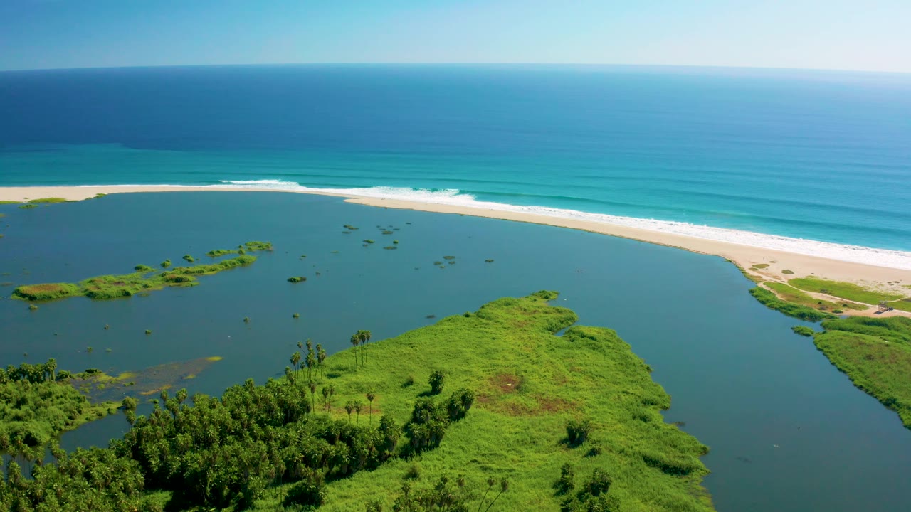 The aerial view over San Jose del Cabo shows small patches of vegetation in the lagoon, a bright sandy beach, layered turquoise ocean water, and no visible people.