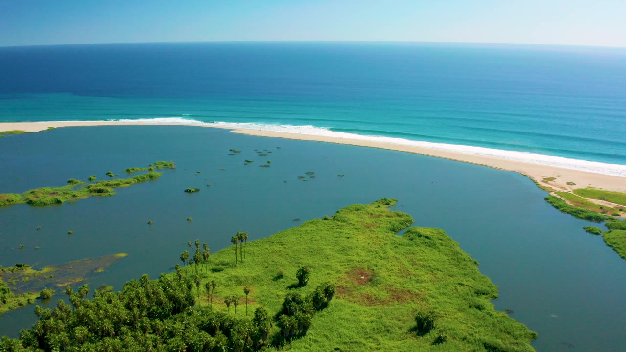 Aerial view in San Jose del Cabo showing green lagoon vegetation in the lower frame, calm blue lagoon water, a pale sandy beach, turquoise ocean, and no visible people.