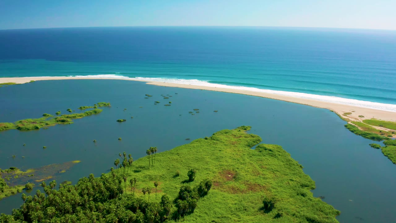 Aerial frame over San Jose del Cabo with the green landmass centered lower in frame, lagoon water wrapping around it, surf breaking along the beach, and no visible people.
