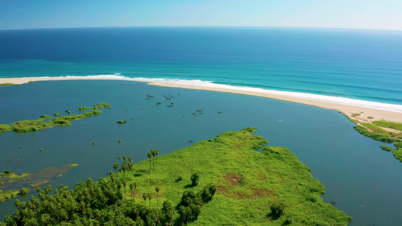 San Jose del Cabo aerial view with small vegetation patches in the lagoon, a bright beach band across the frame, blue-green ocean water, and no visible people.