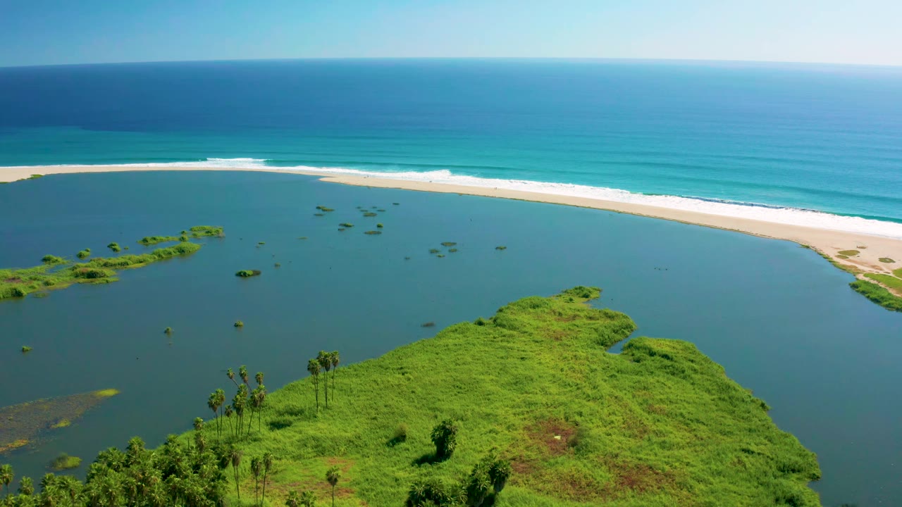 Aerial view in San Jose del Cabo with the green foreground point lower in frame, scattered vegetation in the lagoon, white surf on the sandbar, and no visible people.
