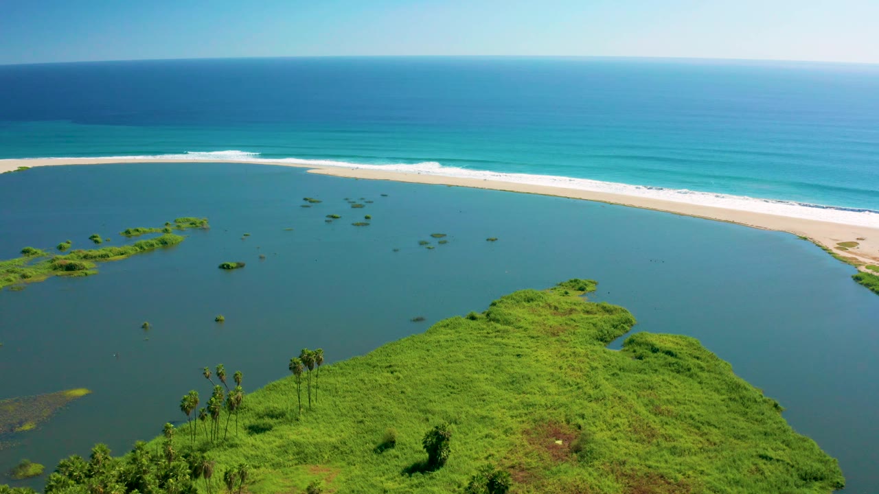 Aerial San Jose del Cabo view with lagoon water filling most of the frame, green vegetation along the lower edge, a pale beach and white surf in back, and no visible people.