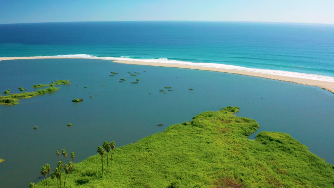 Aerial frame over San Jose del Cabo lagoon with a grassy point and palm trees in the foreground, small green islets in turquoise water, a pale beach, white surf, blue ocean, and no visible people.