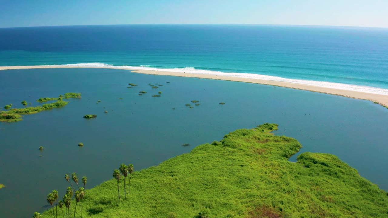 San Jose del Cabo coastal lagoon seen from above with the grassy foreground still prominent, scattered green islets in the water, a long sand beach, white wave line, ocean beyond, and no visible people.