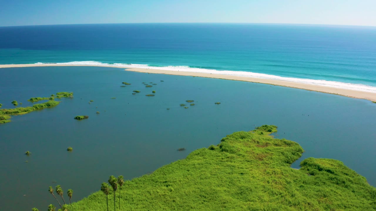 Aerial coastal lagoon with green shoreline, islets, beach, waves, and blue ocean.