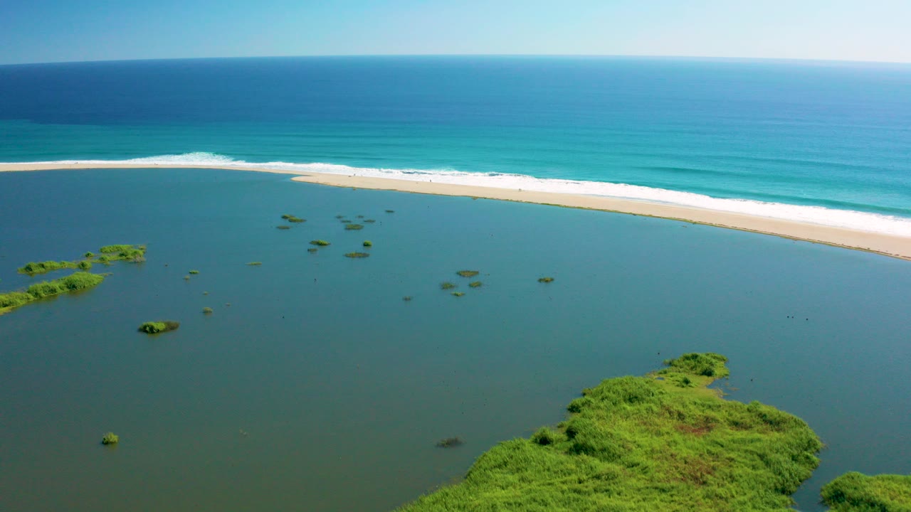 Aerial frame over San Jose Del Cabo showing a calm lagoon with green islets, a pale sandbar, blue ocean waves beyond it, and no visible people.