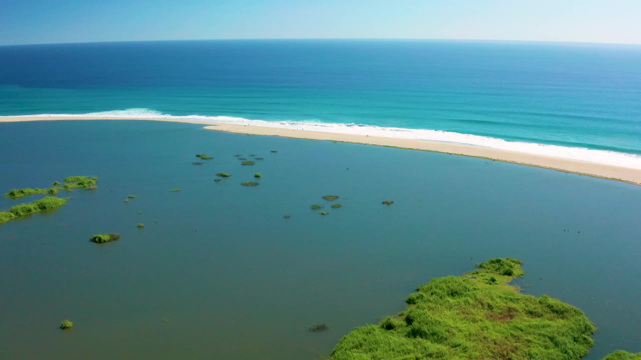 Aerial frame over San Jose Del Cabo with a larger green islet entering the lower right, smaller islets in the lagoon, a bright sandbar, blue ocean, and no visible people.