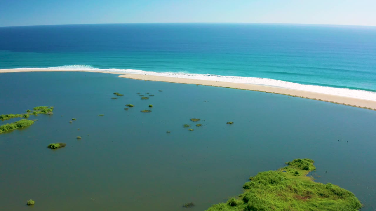 Wide San Jose Del Cabo aerial frame showing open lagoon water, small green islets, foreground vegetation along the bottom, a pale beach strip, ocean waves, and no visible people.