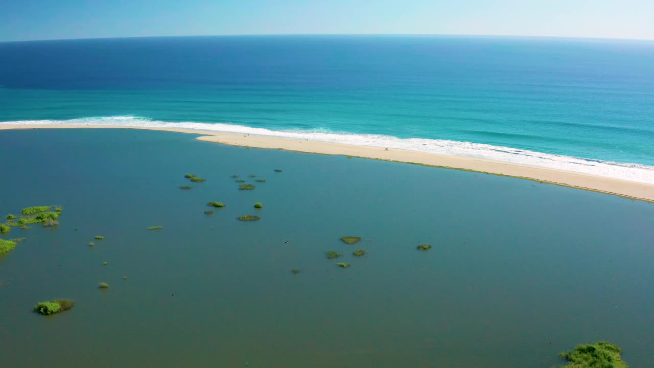 Aerial view in San Jose del Cabo showing small green vegetation patches scattered through the lagoon below a narrow sandbar and blue ocean, with no visible people.