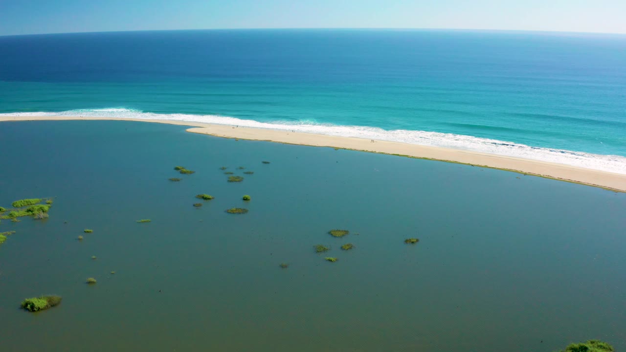 High aerial frame over San Jose del Cabo with the curved sandy beach running across the middle, darker lagoon water below, turquoise ocean above, and no visible people.