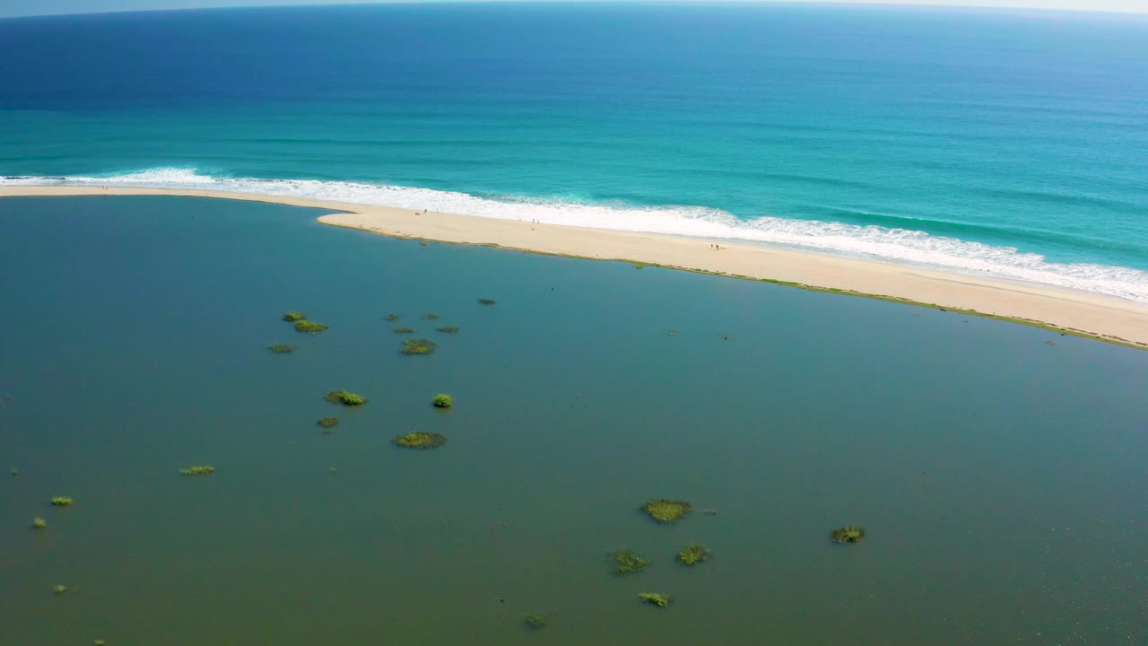 Aerial frame from San Jose del Cabo with white waves advancing along the beach, darker shallow water in the foreground, small reef patches, and no visible people.