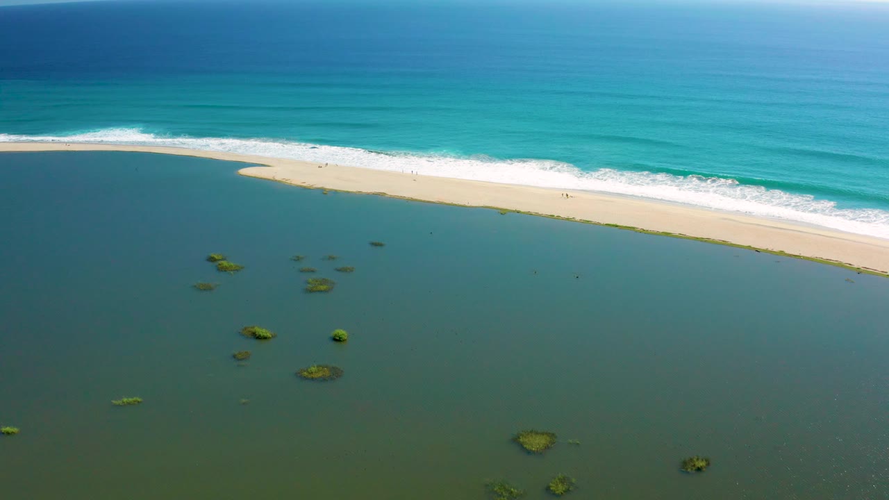 Aerial frame from San Jose del Cabo where shallow green-blue water fills more of the foreground, small reef patches sit below the camera, surf breaks beyond the sand, and no visible people appear.
