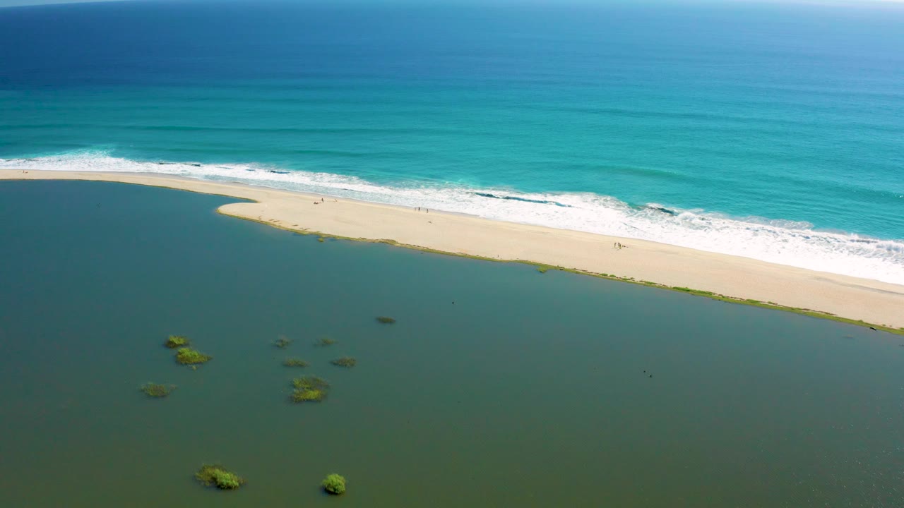 Aerial view over San Jose del Cabo coastline with turquoise water above the surf, a narrow pale beach, and calm darker water below it, no visible people.
