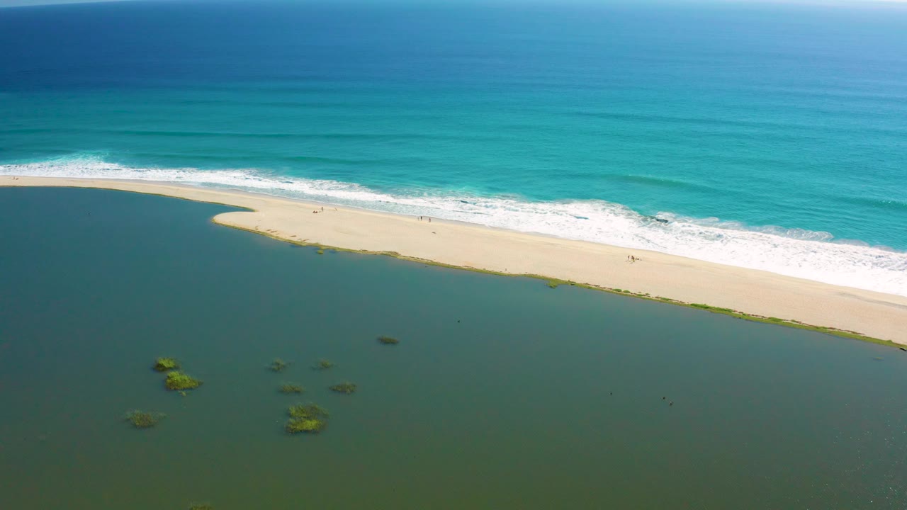 San Jose del Cabo aerial shoreline frame with white surf curving along the beach and scattered green patches in the darker shallow water, no visible people.