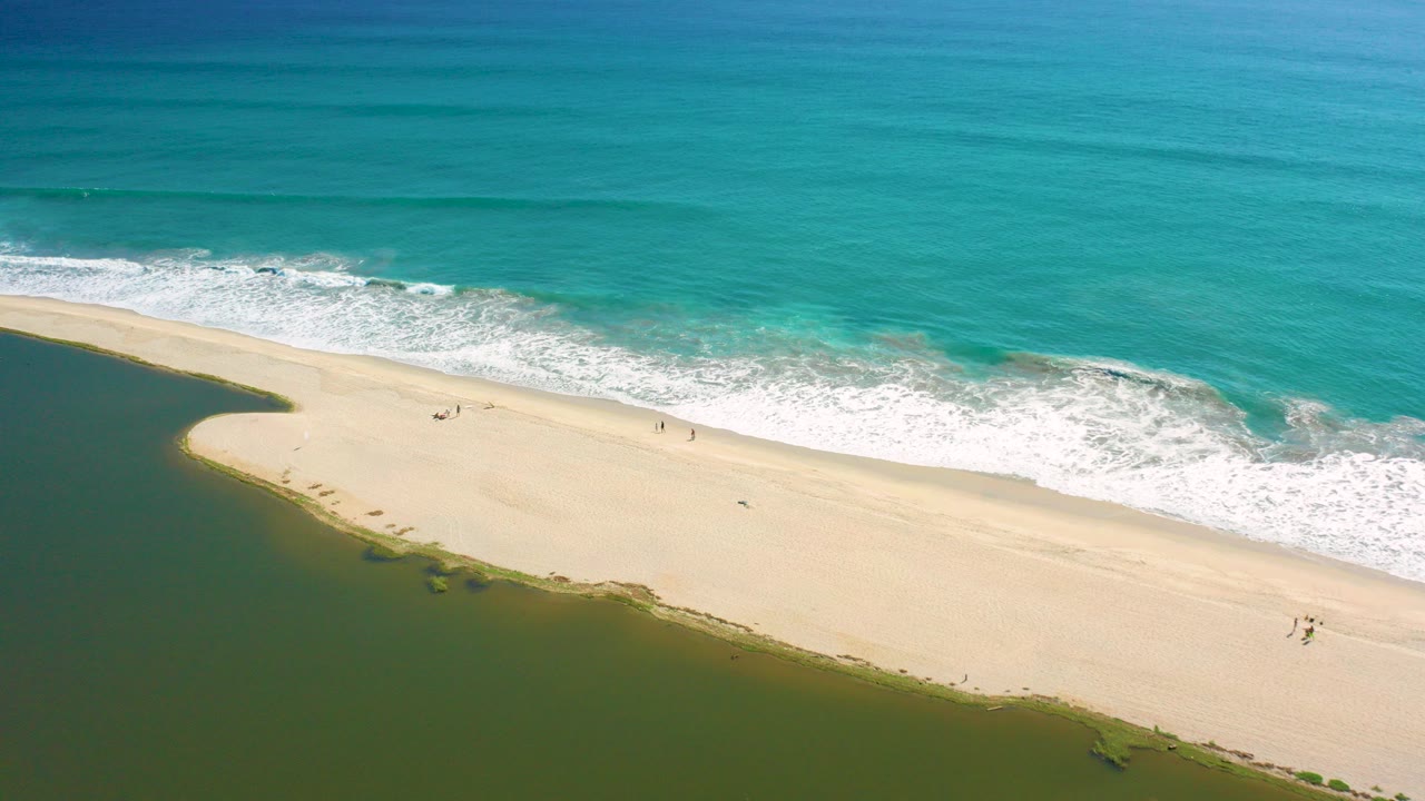 Aerial frame over San Jose del Cabo showing pale sand between turquoise ocean waves and green lagoon water, with tiny walkers visible near the shoreline.