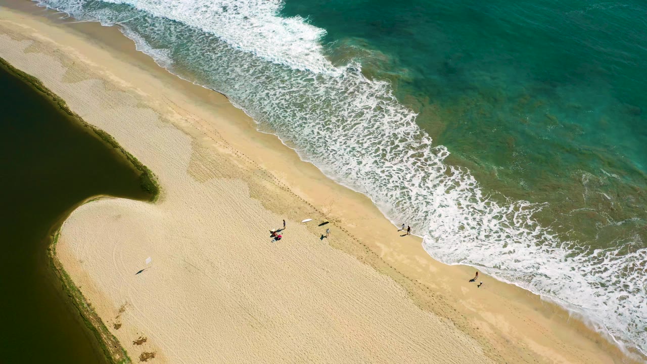 Overhead view in San Jose Del Cabo, Mexico, showing a sandy beach between a dark lagoon and turquoise ocean as white surf reaches small people near the shore.