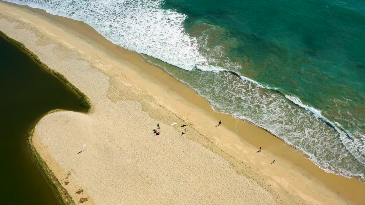 Overhead view of San Jose del Cabo beach with turquoise surf at the top right, a dark inlet at the left, pale sand through the center, and several small beachgoers near the waterline.