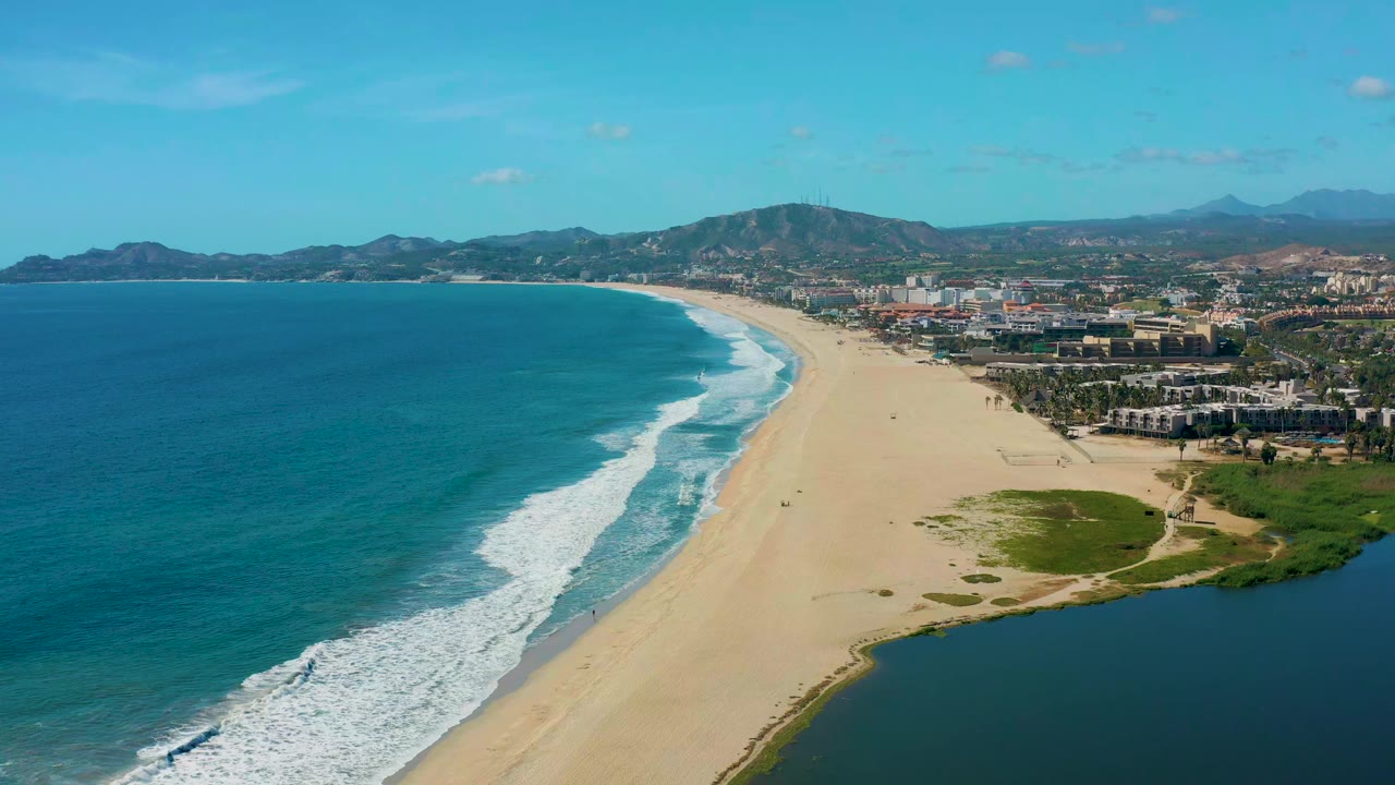 Aerial view in San Jose del Cabo with the surf line slightly farther down the beach, turquoise water at left, pale sand through the center, a green shoreline patch, buildings, hills, and no visible people.