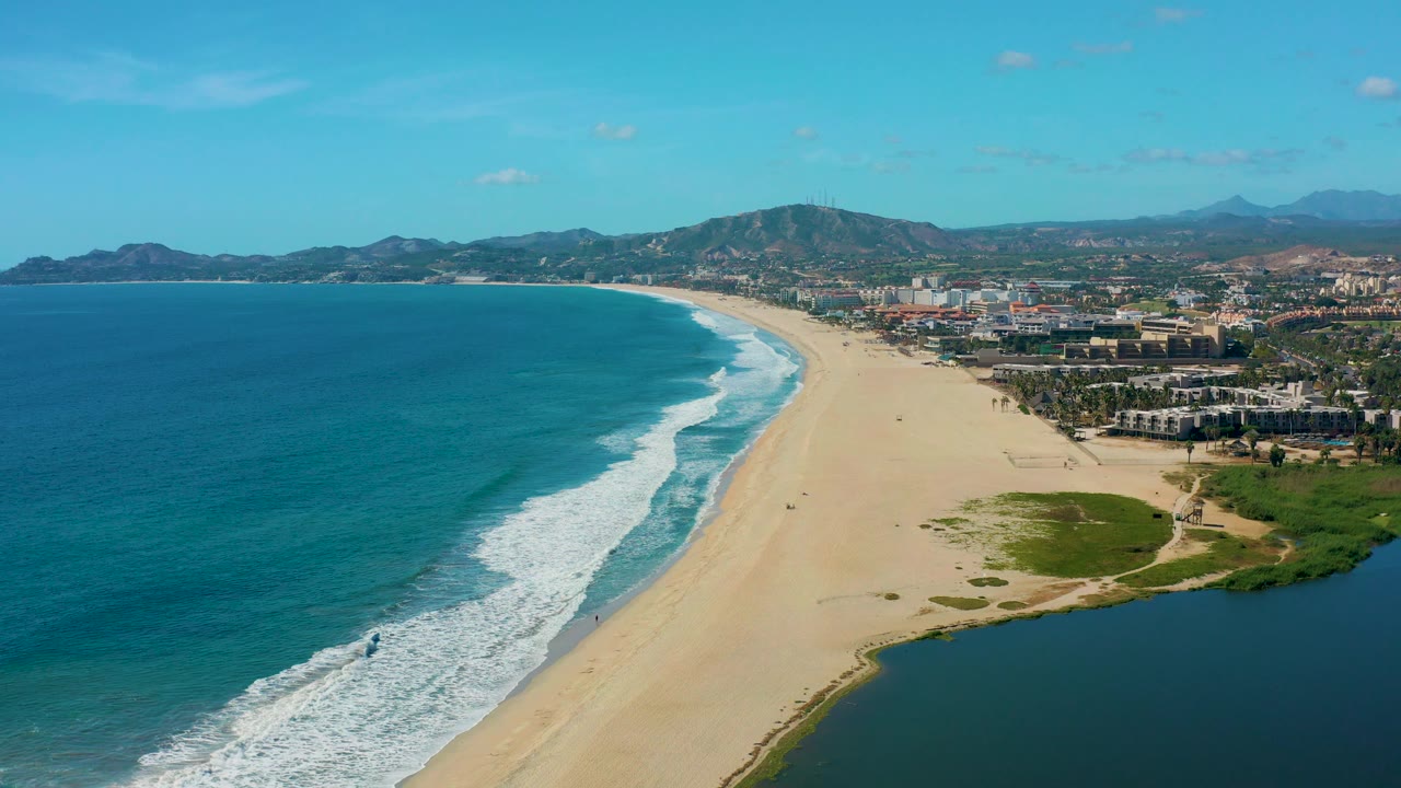 Mid-clip aerial over San Jose del Cabo with broad blue water to the left, surf touching the sand, a dark lagoon or inlet at lower right, coastal development beyond the beach, hills, and no visible people.