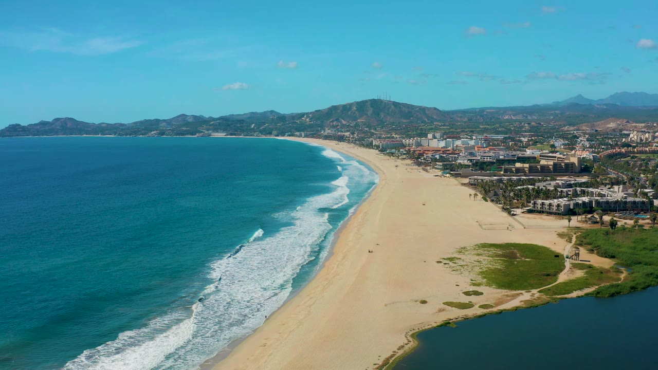 San Jose Del Cabo aerial view one second in, with white wave bands closer to the beach, pale sand running through the center, green lagoon margins at lower right, and no visible people.