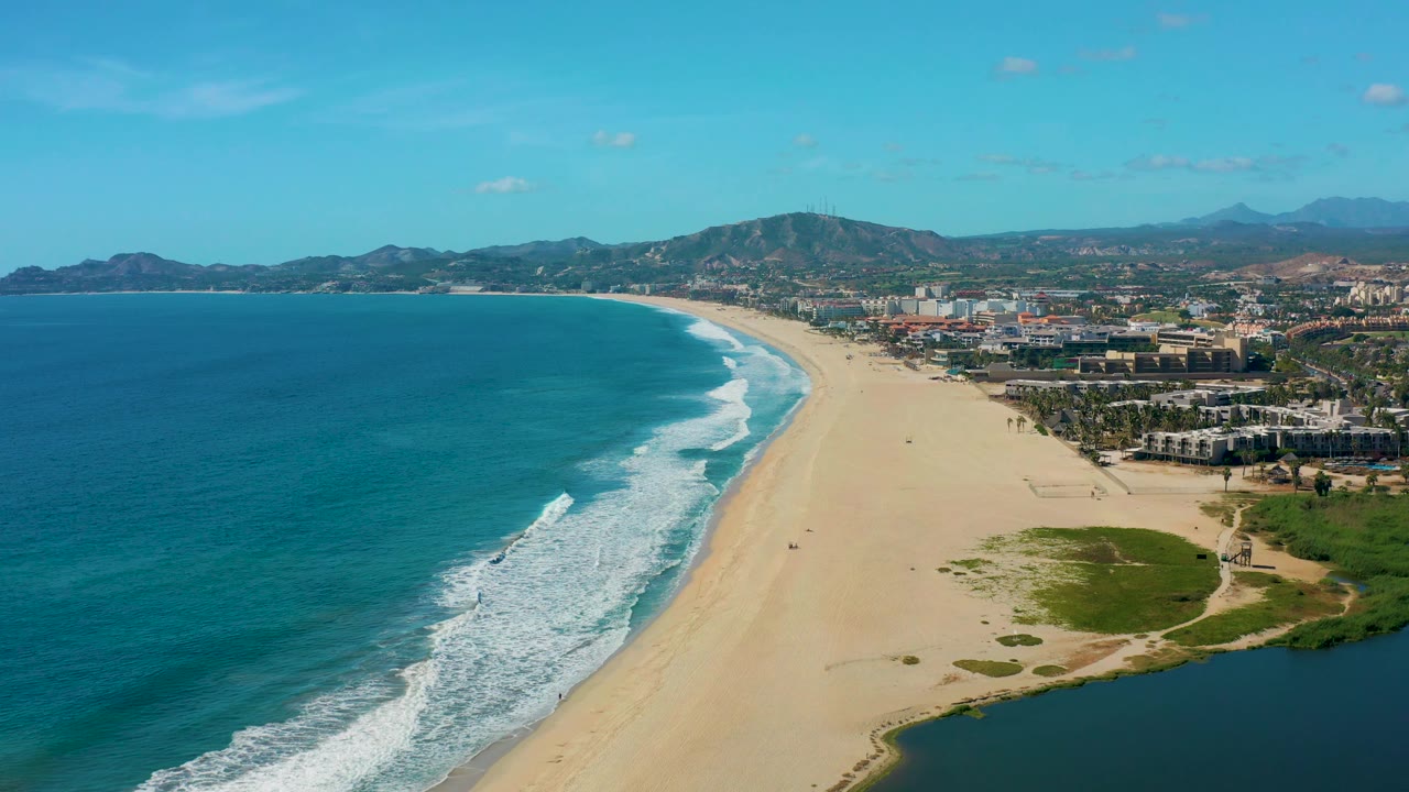 At 0:02 the aerial composition keeps the ocean sweeping in from the left, the beach tapering into the distance, a darker lagoon corner at lower right, and no visible people in San Jose Del Cabo.
