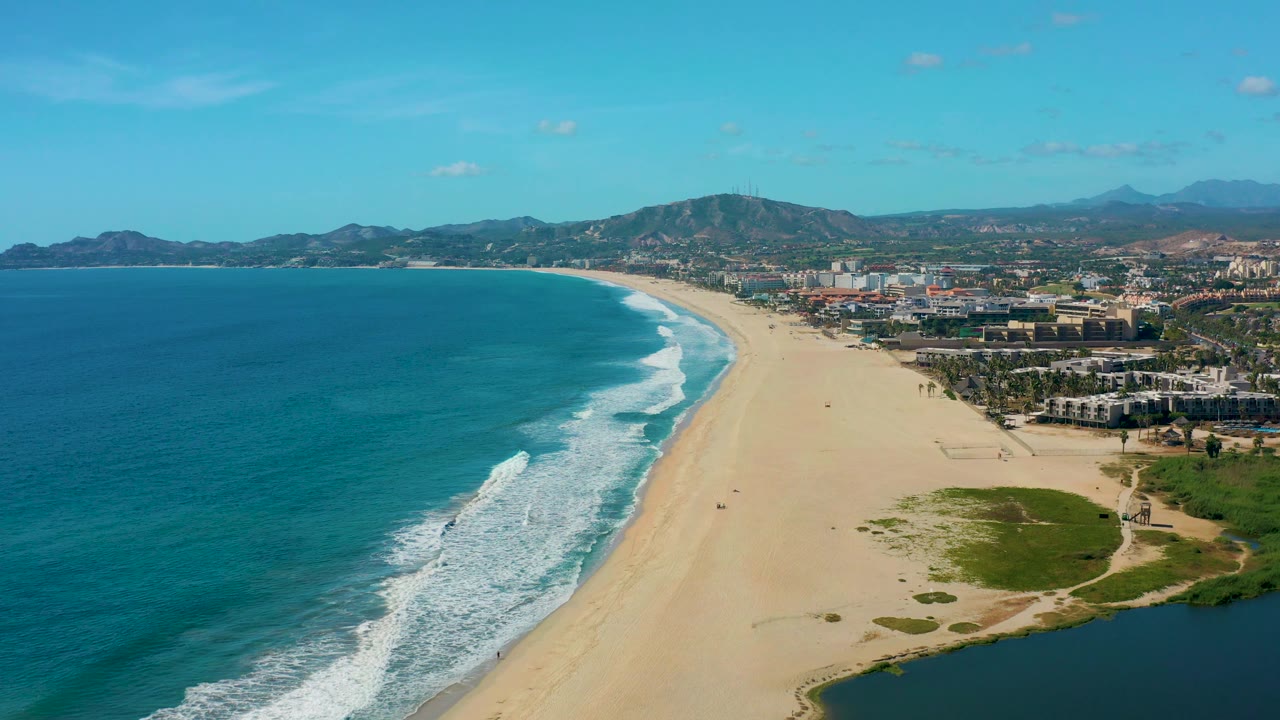 Mid-clip frame over San Jose Del Cabo showing the surf line slightly farther along the sand, a green lagoon area in the lower right foreground, distant buildings, mountains, and no visible people.