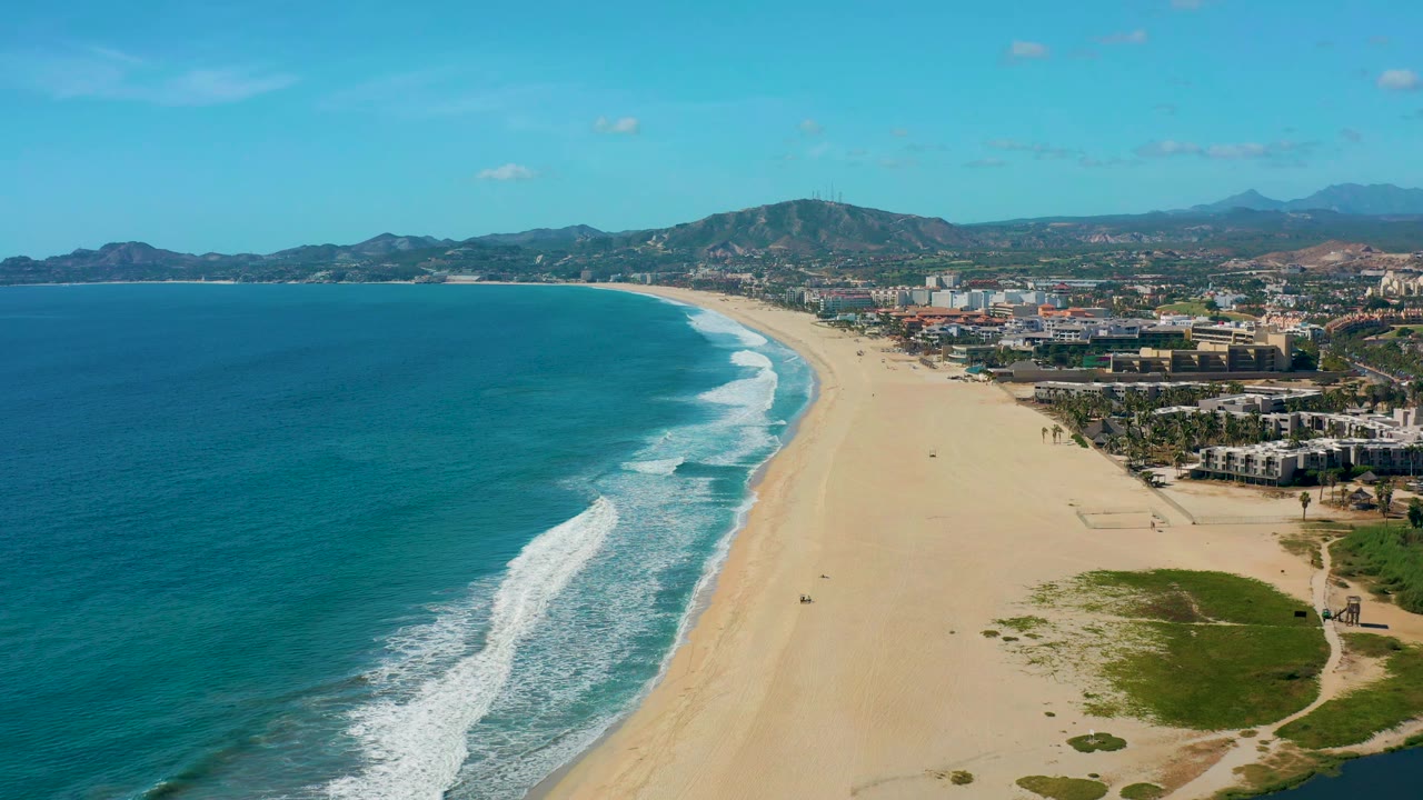 Aerial San Jose Del Cabo coastline frame showing surf bands curving toward the sand, clustered coastal buildings on the right, mountains in the distance, and no visible people.