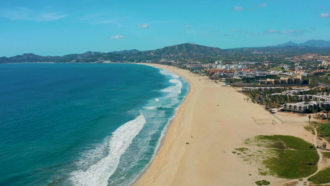 San Jose Del Cabo aerial view with blue ocean filling the left side, white wash along the beach, coastal buildings and roads on the right, mountains beyond, and no visible people.