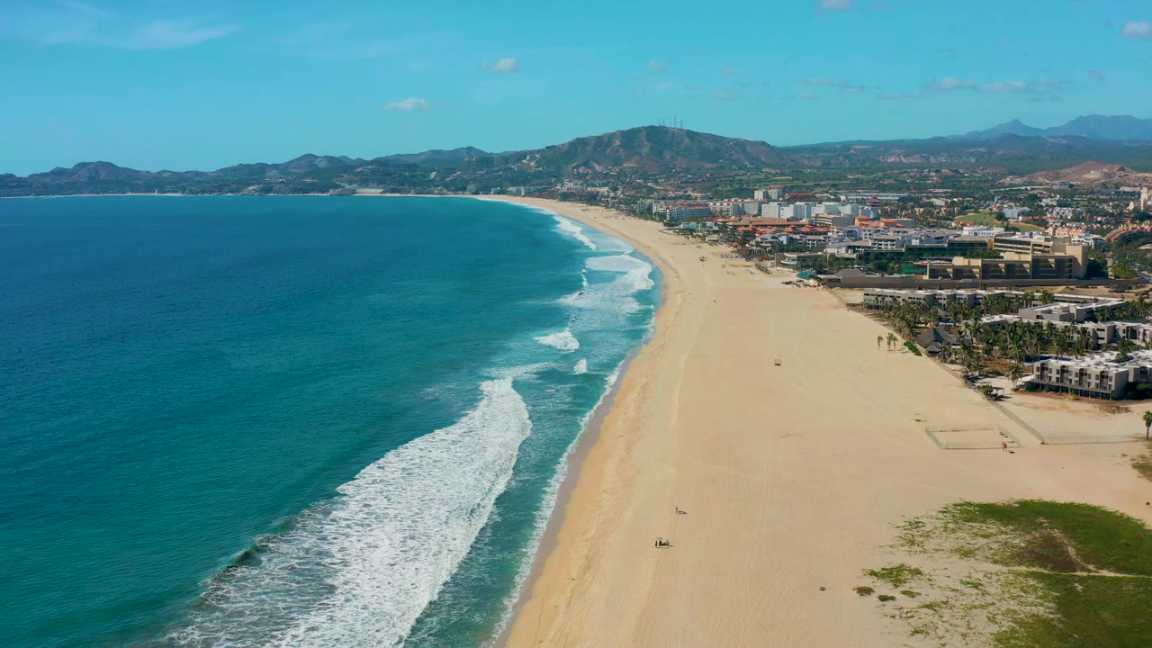 Drone view of white surf rolling toward a long sandy beach with buildings and mountains behind.