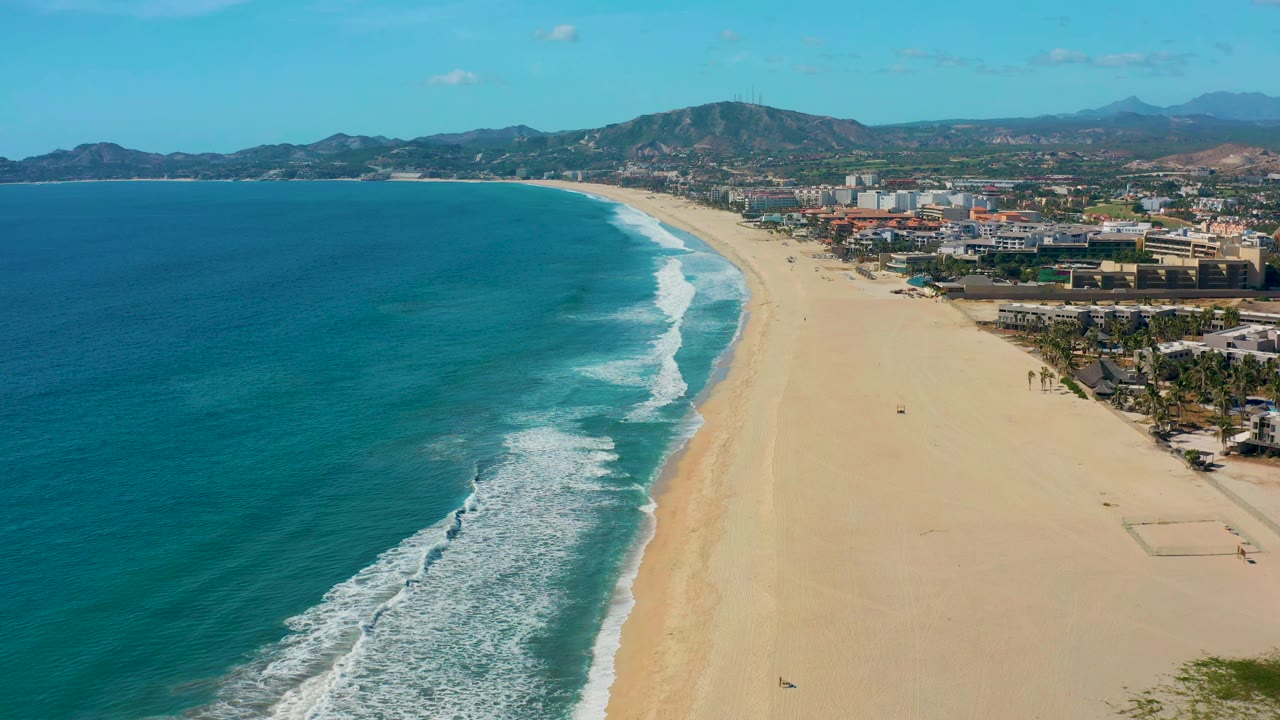 Curving surf line on a wide sandy beach with town and hills beyond.