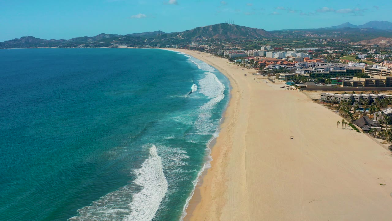 Opening aerial frame over San Jose del Cabo with turquoise ocean on the left, white surf curving onto the sand, beachfront buildings to the right, distant mountains, and no visible people.