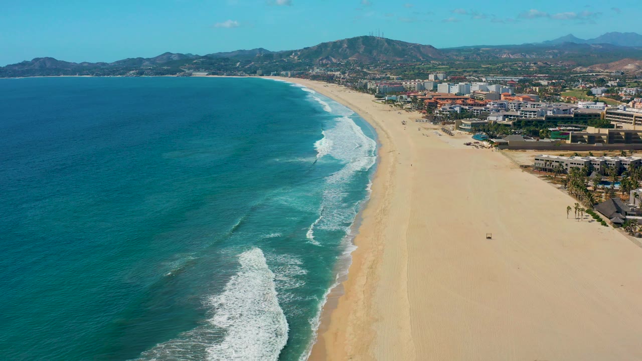 Aerial beach view with surf reaching the shore and mountains behind coastal buildings.