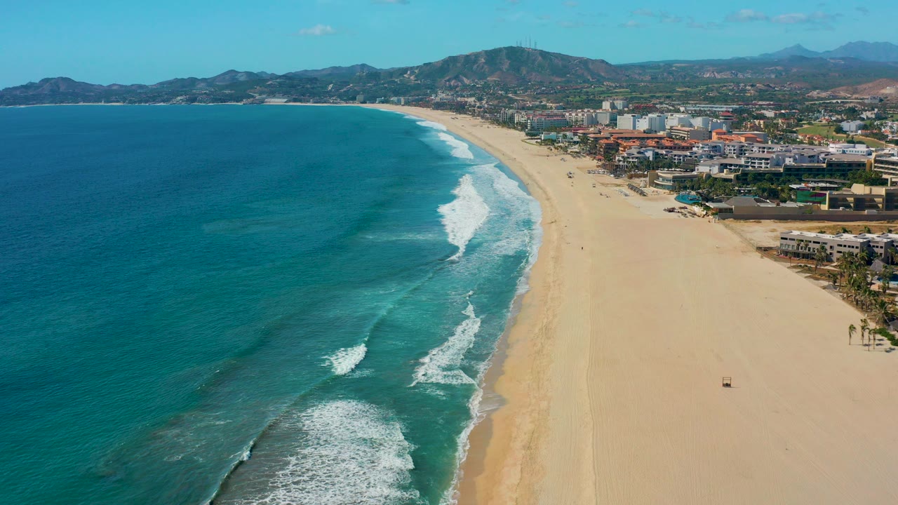 Aerial view over San Jose del Cabo with turquoise ocean on the left, wide sandy beach on the right, coastal buildings beyond the shore, distant mountains, and no visible people.