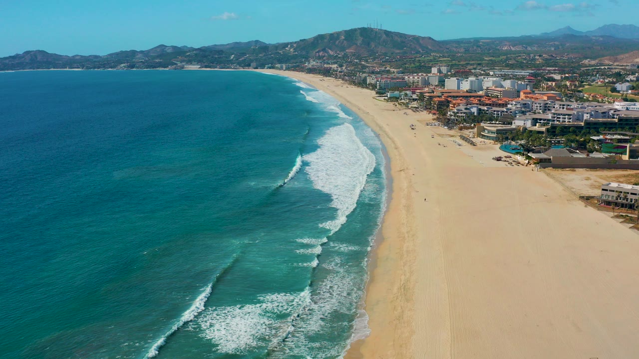 Aerial frame over San Jose del Cabo with blue ocean on the left, white surf approaching a wide sandy beach, beachfront buildings on the right, mountains beyond, and no visible people.