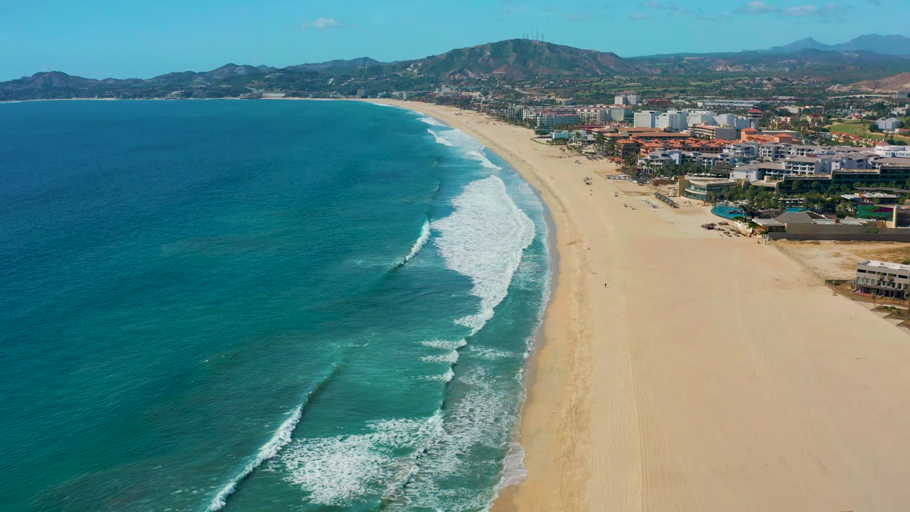 Ocean surf breaks along a long sandy shoreline with buildings and mountains inland.