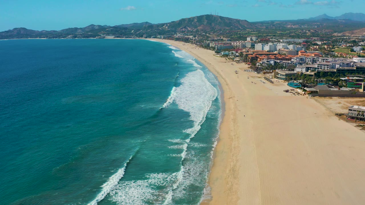San Jose del Cabo coastline from above, with turquoise water spread across the left, a stronger white breaker near the center shore, beachfront development to the right, and no visible people.